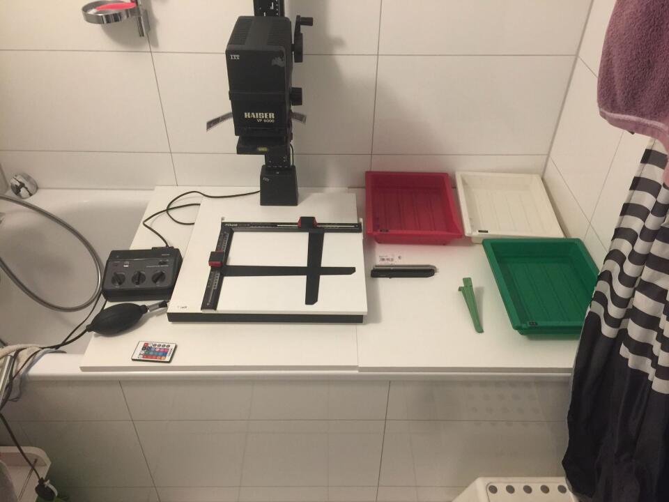 A photo of a bathtub, largely covered by two shelf boards. On top: a photographic enlarger, a timer, three trays with chemicals.