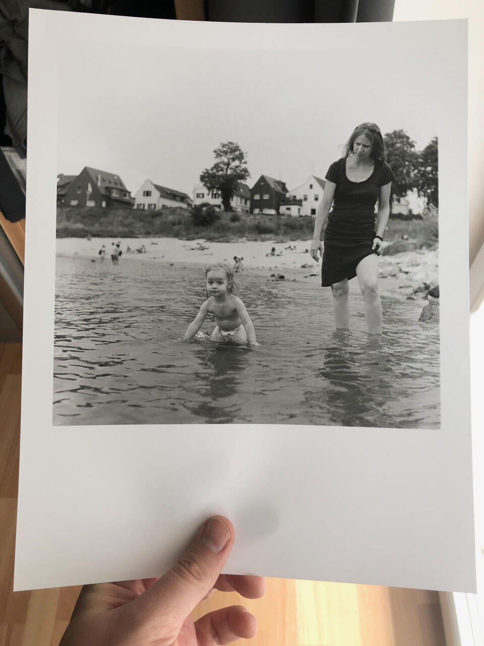 A child waist-high in a river. A woman next to it. Both facing the camera, but not looking into it. A beach and houses in the background.