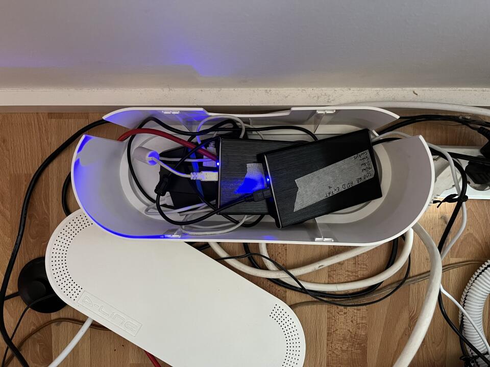 The Raspberry Pi in a case, buried under two HDDs and a bunch of cables. All in a cable box – lid next to it – on the floor, photographed from above.
